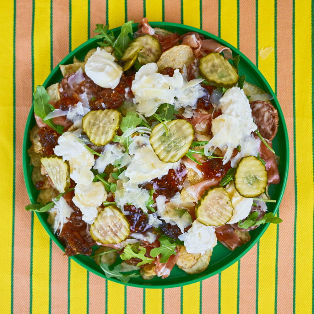 Charcuterie nachos on a green plate with an orange and yellow striped tablecloth.
