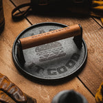 Cast iron burger press with wooden handle on a wooden surface, surrounded by leather goods.