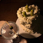 Decorative cake with olive candles next to a cocktail glass on a wooden surface
