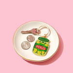 Keychain shaped like a pickle jar on a white dish with coins and keys against a pink background