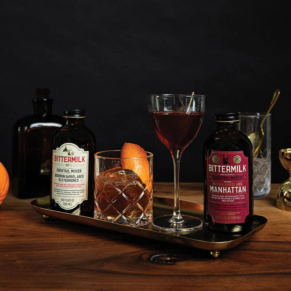 Bittermilk cocktail bottles and glasses on a wooden tray with a dark background