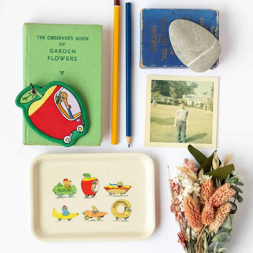 Collection of items including a green book, colorful patch, pencil, photo, and decorative Richard Scarry tray on a white surface.
