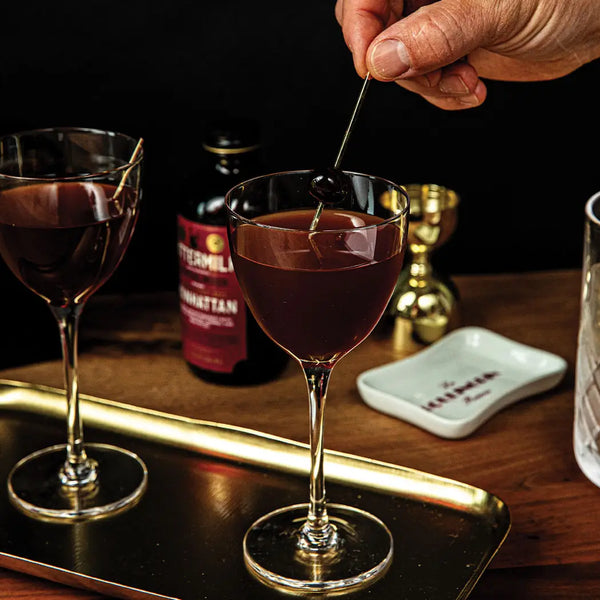 Two cocktail glasses with a dark liquid on a gold colored tray, with a bottle of Bittermilk Cocktail mixer blurred in the background.