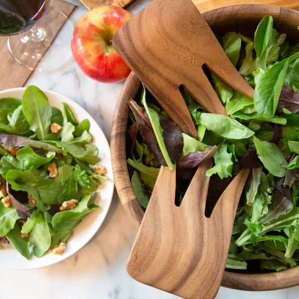 Wooden salad servers in a bowl of salad with an apple and glass of wine in the background.