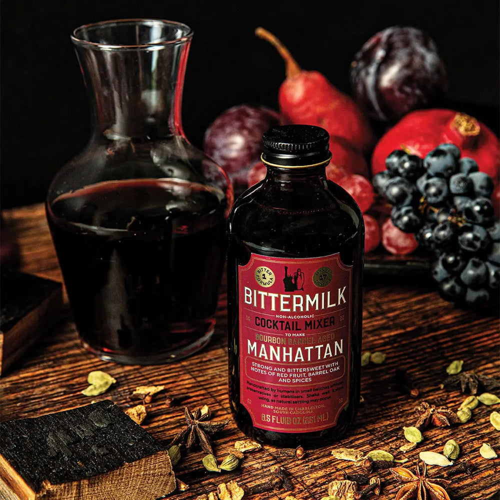 Bittermilk cocktail mixer bottle with a glass carafe on a wooden surface surrounded by fruits and spices.