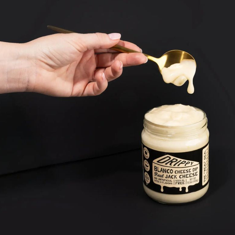 Hand holding a spoon with cheese over a jar of Drippy Blanco Cheese Dip on a black background