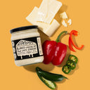 Jar of Drippy Blanco Cheese Dip surrounded by cheese, peppers, and jalapeños on a yellow background.