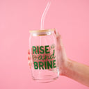 Hand holding a glass can with straw and 'Rise and Brine' text on a pink background