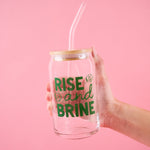 Hand holding a glass can with straw and 'Rise and Brine' text on a pink background
