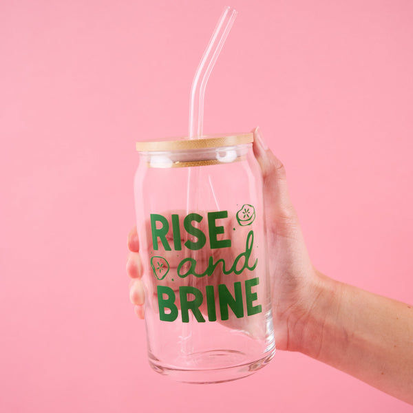 Hand holding a glass can with straw and 'Rise and Brine' text on a pink background