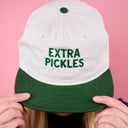 Person wearing a white and green cap with 'Extra Pickles Please' text against a pink background