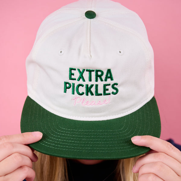 Person wearing a white and green cap with 'Extra Pickles Please' text against a pink background