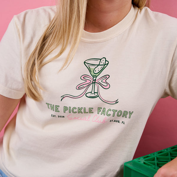 Person wearing a white t-shirt with 'The Pickle Factory Social Club' logo against a pink background