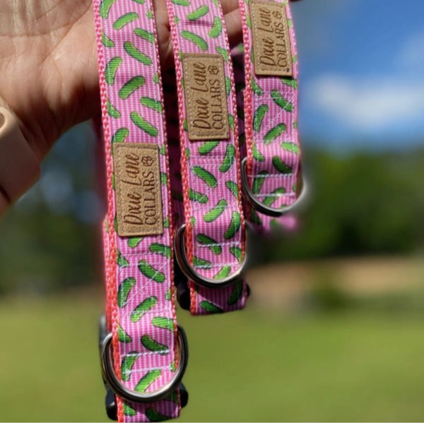 Three different sizes of pink dog collars with a pickle pattern in front of a outdoor landscape