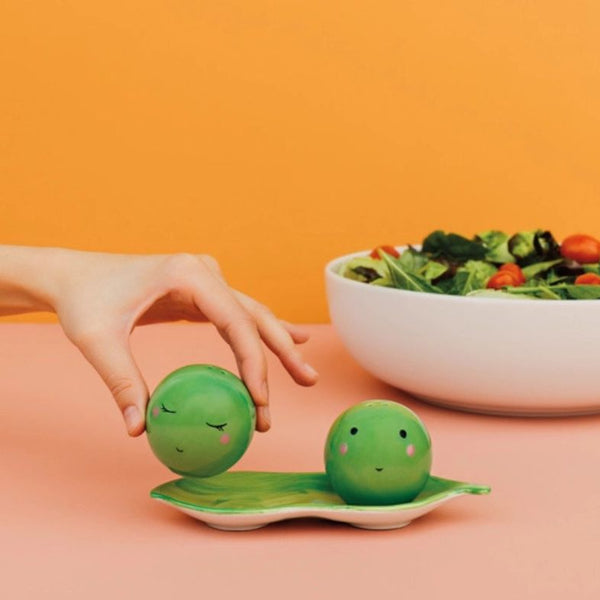 Person holding a green pea-shaped salt and pepper shaker with a face on it with an orange background.