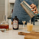 Person making a cocktail using Bittermilk cocktail mixer on a kitchen counter.
