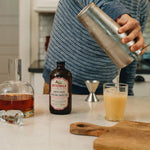 Person making a cocktail using Bittermilk cocktail mixer on a kitchen counter.
