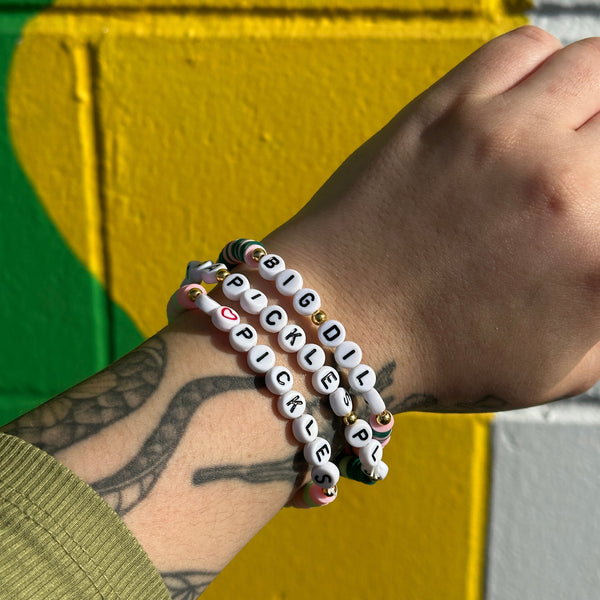 A person's wrist wearing 3 pickle-themed friendship bracelets against against a colorful background.