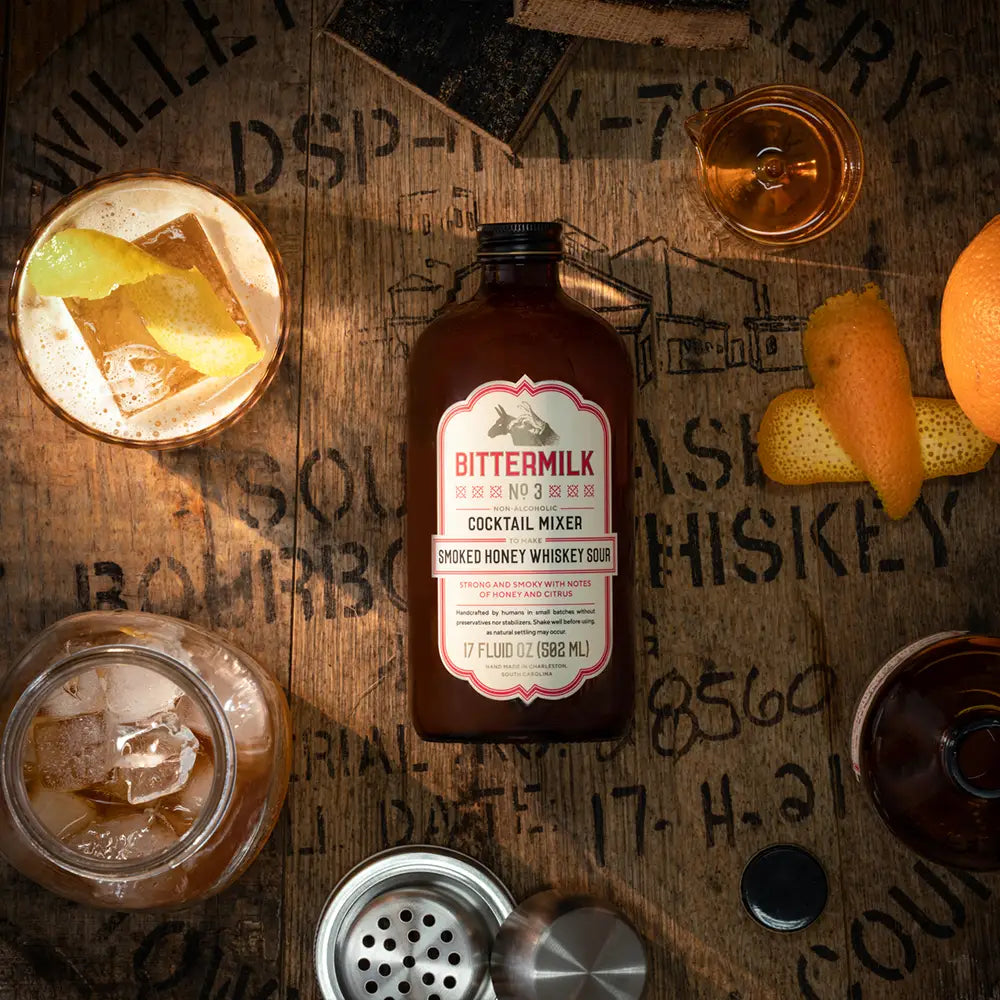 Bittermilk cocktail mixer bottle on a wooden surface surrounded by cocktail ingredients.